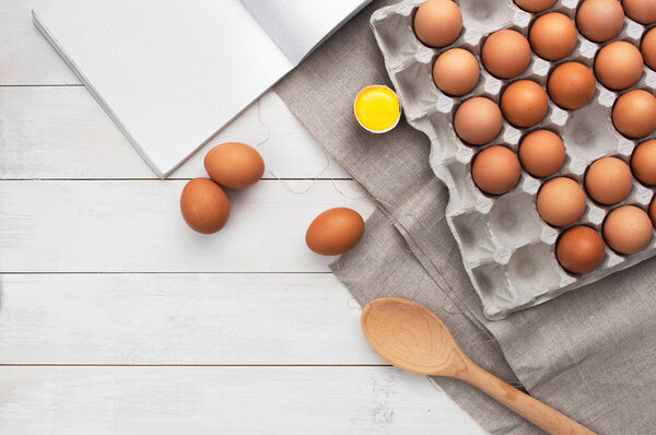 Top view of chicken eggs, culinary notebook and spoon on white wooden table with copy space.
