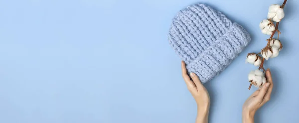 Piatto laici alla moda blu maglia cappello invernale in mani femminili fiori di cotone su sfondo blu vista dall'alto. Elegante cappello di lana concetto di accessori invernali per il freddo. Pubblicità shopping inverno vendita — Foto Stock