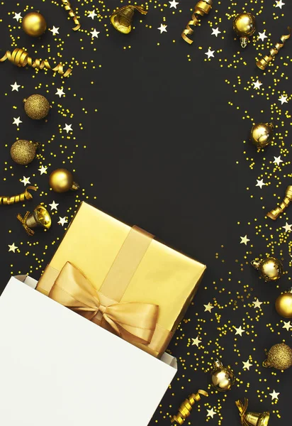 New Year or Christmas composition. Golden gift with ribbon from white paper bag, Christmas balls, confetti stars on black background. Flat lay top view. Golden decorations, party holiday concept 2020 — Stock Photo, Image