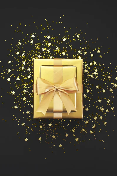 Regalo dorado con cinta, estrellas de confeti sobre fondo negro. Piso tendido, vista superior, espacio para copiar. Decoraciones doradas, fiesta, cumpleaños, concepto de vacaciones. Caja de regalo, sorpresa. Año Nuevo o composición de Navidad — Foto de Stock