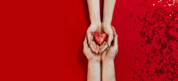 Kobieta i mężczyzna ręce trzymając czerwony kształt serca Makaron ciasto i konfetti na czerwonym tle topview Flat lay. Kreatywna kompozycja na Walentynki koncepcja miłości urodziny prezent dzień matki wakacje — Zdjęcie stockowe