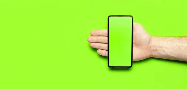 Male hands hold a modern black smartphone with green blank screen on neon green background flat lay top view. Modern technology, phone, gadget in hands, touch screen, template for your design. Mockup