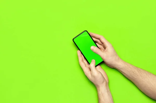 Чоловічі руки тримають сучасний чорний смартфон із зеленим порожнім екраном на неоновому зеленому фоні з плоским видом зверху. Сучасні технології, телефон, гаджет в руках, сенсорний екран, шаблон вашого дизайну. Мокап — стокове фото