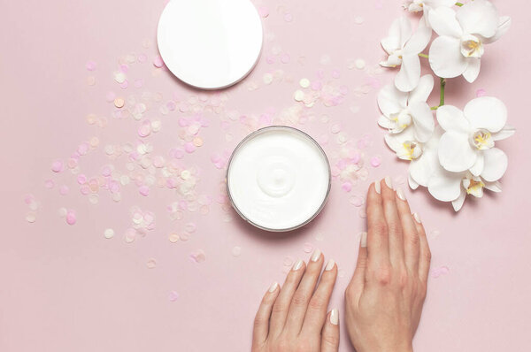 Young woman holding a opened container with cosmetic cream lotion body milk White Phalaenopsis orchid flowers festive confetti on pink background Flat lay top view minimalism style Beauty concept....