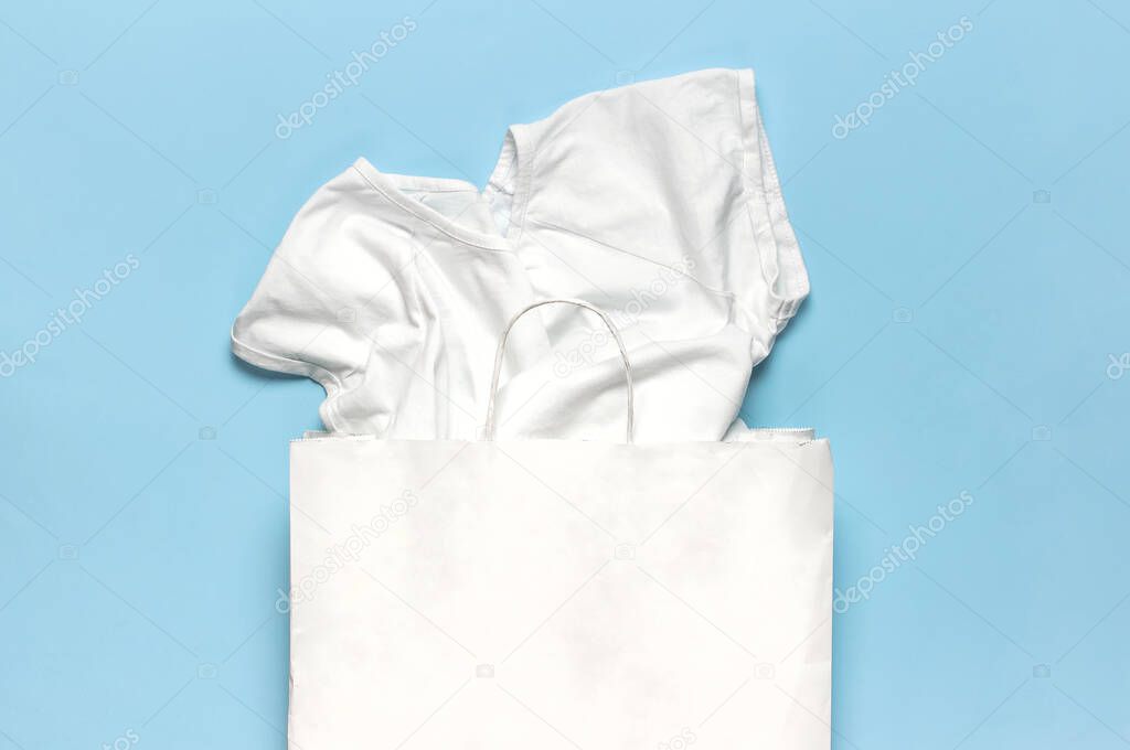 White shirt, paper bag on blue background top view flat lay. Fashion discounts shopping sale store promo design. Women's clothing, basic t-shirt. Place for text.