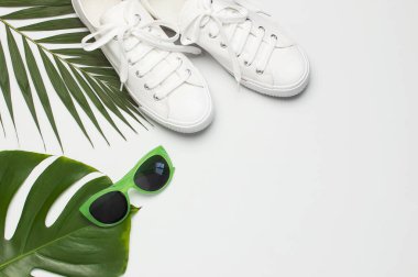 Beyaz kadın moda ayakkabıları, modaya uygun güneş gözlükleri, açık gri arka planda tropikal yapraklar. Düz görünüm kopyalama alanı yatıyordu. Kadın ayakkabıları, aksesuarlar. Şık beyaz ayakkabılar. Yaratıcı yaz konsepti