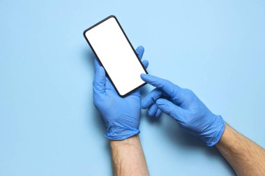 Mavi eldivenli erkek eller mavi arka planda beyaz beyaz ekranlı akıllı telefonu tutuyor. Virüs sırasında bakterilere karşı güvenlik koruması, Coronavirus 2019-nCoV. Telefonlu el doktoru..