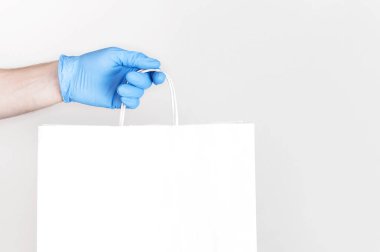 Karantina sırasında mal teslimatı. Mavi eldivenli genç adam beyaz arka planda beyaz kağıt torba tutuyor. Coronavirus 2019-NCov 'a karşı salgın halinde teslim.