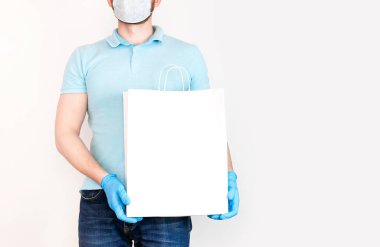 Tıbbi maskeli ve mavi eldivenli sakallı genç adam beyaz arka planda beyaz kağıt torba tutuyor. 2019 ncov Kontaksız dağıtım virüsü karantinada mal teslimi kavramı.
