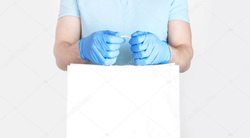 Concept of delivery of goods during quarantine. Young man in blue gloves holds white paper bag in his hands on white background. Delivery against Coronavirus 2019-nCov in pandemic Contactless delivery.