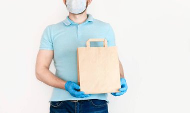 Tıbbi maskeli ve mavi eldivenli sakallı genç adam beyaz arka planda el işi kağıt torba tutuyor. 2019 ncov Kontaksız dağıtım virüsü karantinada mal teslimi kavramı.