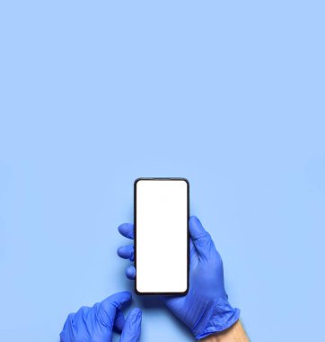 Mavi eldivenli erkek eller mavi arka planda beyaz beyaz ekranlı akıllı telefonu tutuyor. Bir salgın sırasında sağlığın korunması 2019 nCoV Eller Sağlık Görevlisi Telefonlu gönüllü doktor.
