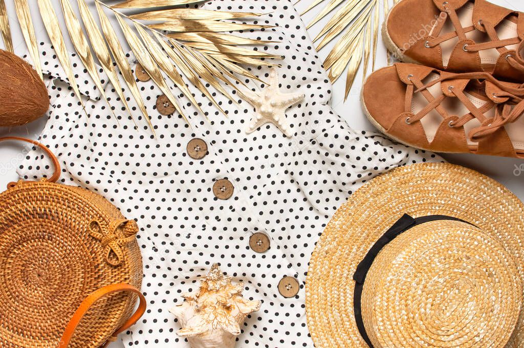 Summer womens white dress in black peas rattan woven bag brown sandals straw hat golden palm leaf shells starfish on light background. Flat lay top view. Womens beach fashion, travel vacation
