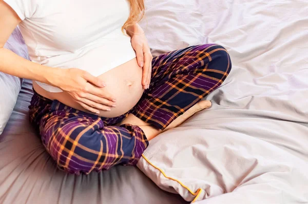 Konzept der Erwartung des Kindes, Schwangerschaft und Mutterschaft. Die junge schwangere Frau sitzt zu Hause im Bett und berührt ihren Bauch mit den Händen. Glückliche Schwangerschaft, Warten auf die Geburt eines Babys — Stockfoto