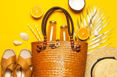 Yaz arkaplanı plaj aksesuarları. Plaj hasır hasır hasır kadın eko çantası sandaletleri altın tropikal yaprak Hindistan cevizi portakalı, meyve suyu, pipet, sarı arka planda deniz yıldızı. Düz üst görünüm.