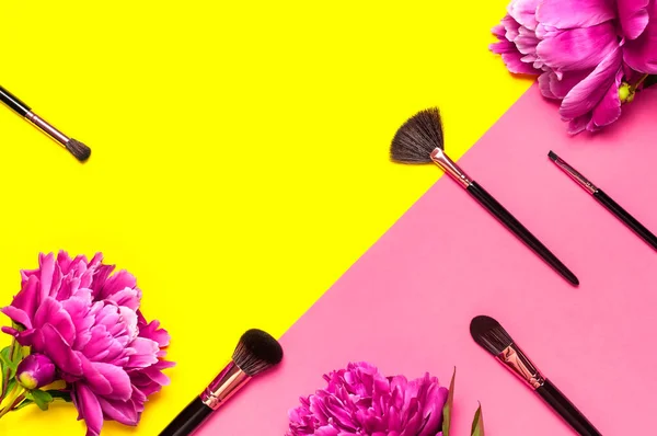 Professional Different makeup brushes with pink spring peonies flowers on yellow pink background flat lay top view copy space. Beauty product, makeup, women's accessory, fashion. Cosmetic makeup Set.