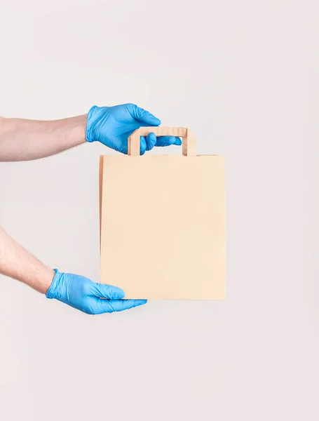Delivery against Coronavirus 2019-nCov in pandemic Contactless delivery. Male hand in blue medical gloves holds craft paper bag on white background. Concept of delivery of goods food during quarantine.