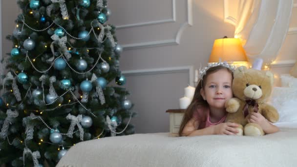 Mulher está em uma cama com um urso de pelúcia perto de uma árvore de Natal — Vídeo de Stock