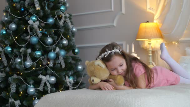 Mulher está em uma cama com um urso de pelúcia perto de uma árvore de Natal — Vídeo de Stock