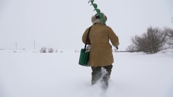 Un pescador camina sobre un lago cubierto de nieve en busca de un buen lugar de pesca . — Vídeo de stock