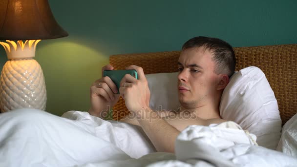 Homem está deitado na cama e está usando o aplicativo em seu smartphone . — Vídeo de Stock