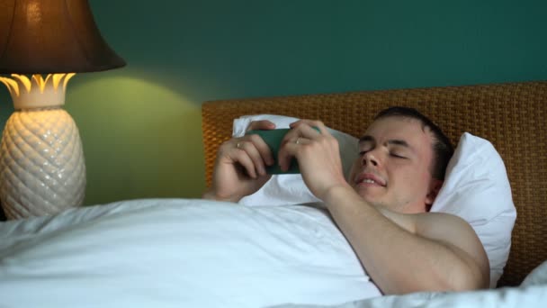 Hombre está acostado en la cama y está utilizando la aplicación en su teléfono inteligente . — Vídeos de Stock