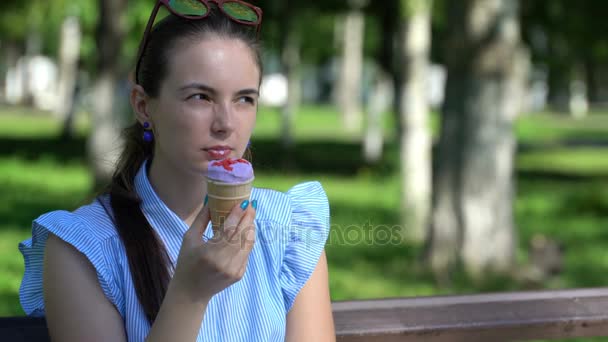 Menina comendo um sorvete sentado em um banco de parque . — Vídeo de Stock