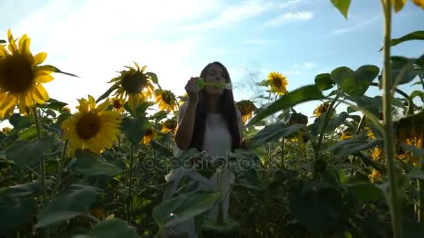 Vacker ung flicka promenader i det gula fältet av solrosor och blåser såpbubblor. — Stockvideo