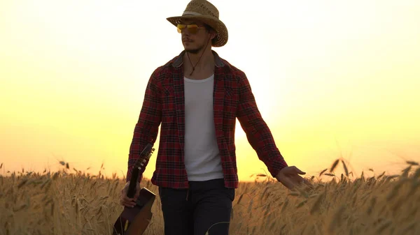 Jovem caminha no campo de trigo dourado nos raios do pôr do sol e toca guitarra . — Fotografia de Stock