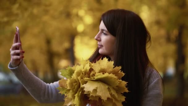 Mädchen spaziert im Herbst durch einen Park und macht ein Selfie mit einem Strauß orangefarbener Blätter — Stockvideo