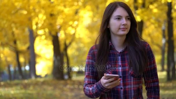 Девушка гуляет в парке осенью и использует приложение на своем смартфоне — стоковое видео