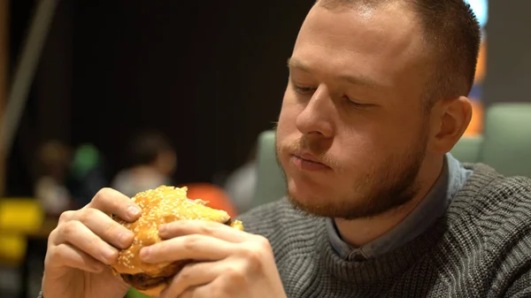 Joven hipster chico comer hamburguesa sentado en la cafetería — Foto de Stock