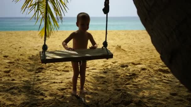 Niño de dos años columpios en un columpio en la playa cerca del océano . — Vídeo de stock