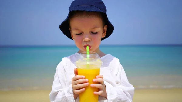Boy of two years drinking mango fresh on the beach on the background of the ocean. — Stock Photo, Image