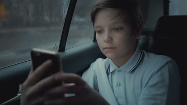 Triste, malheureux jeune garçon chevauchant en voiture à travers la ville pendant la journée de pluie, en utilisant les réseaux sociaux sur son smartphone — Video