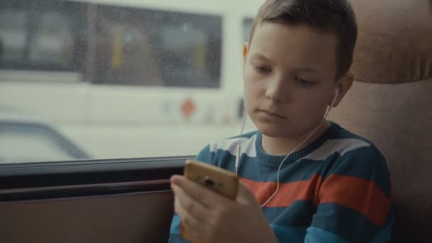 Primer plano de un niño viajando en autobús a través de la ciudad. Él utiliza la red social en su teléfono inteligente y escucha música . — Vídeo de stock