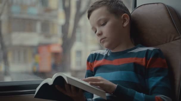 Närbild av en ung skolpojke som reser med buss genom staden och läser en bok. — Stockvideo