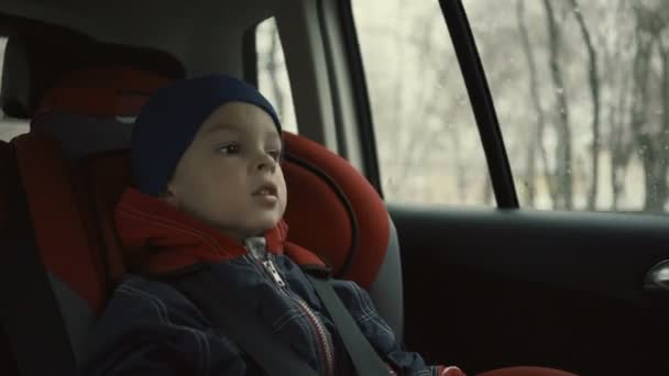 Chico monta en el coche en el asiento del niño y mirando por la ventana en un día lluvioso . — Vídeos de Stock