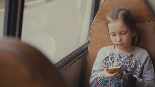 Joven, hermosa pasajera con bolso escolar en el autobús escolar en movimiento utilizando la red social en su teléfono inteligente y mirando por la ventana . — Vídeos de Stock