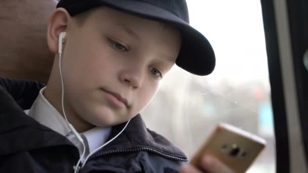 Primer plano de un niño viajando en autobús a través de la ciudad. Él usa un teléfono inteligente y escucha música . — Vídeos de Stock