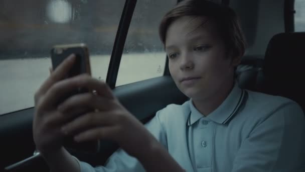 Triste, malheureux jeune garçon chevauchant en voiture à travers la ville pendant la journée de pluie, en utilisant les réseaux sociaux sur son smartphone — Video
