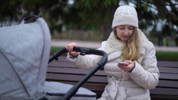 Mère heureuse avec smartphone en utilisant l'application. Elle est assise sur le banc, bébé dormant dans un landau — Video