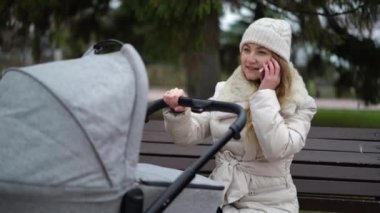 Genç anne, bebek arabasıyla sonbahar parkında yürüyor ve telefonda konuşuyor.