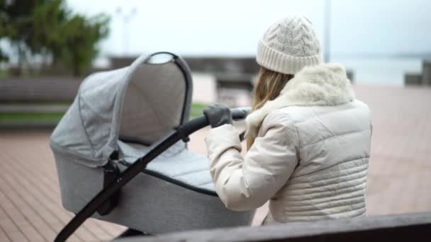 Ung mor med nyfött barn utomhus. Hon sitter på bänken och babyn sover i barnvagn. — Stockvideo