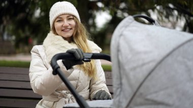 Dışarıda yeni doğmuş çocuğu olan genç bir anne. Bebek arabasında uyurken bankta oturuyor.