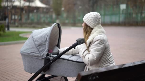 Ung mor med nyfött barn utomhus. Hon sitter på bänken och babyn sover i barnvagn. — Stockvideo