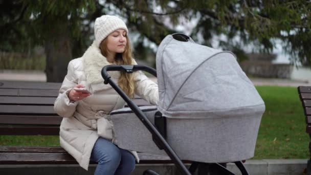 Glückliche Mutter mit Smartphone per App. sie sitzt auf der Bank, Baby schläft im Kinderwagen — Stockvideo