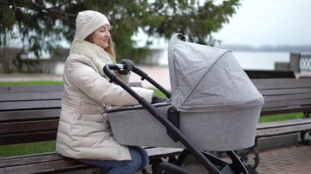 Jovem mãe com filho recém-nascido ao ar livre. Ela sentada no banco com o bebê dormindo em carrinho — Vídeo de Stock