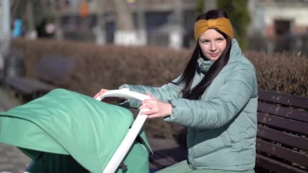 Jeune mère avec nouveau-né en plein air. Elle est assise sur le banc avec bébé dormant dans un landau — Video