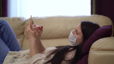 Karantina sırasında evde tıbbi maskeli bir kadın akıllı telefon kullanıyor..